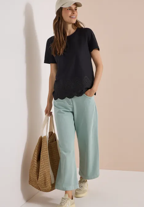 Vrouw draagt zwart T-shirt met golfrand, mintgroene wijde broek, strooien tas, beige sneakers en pet.
