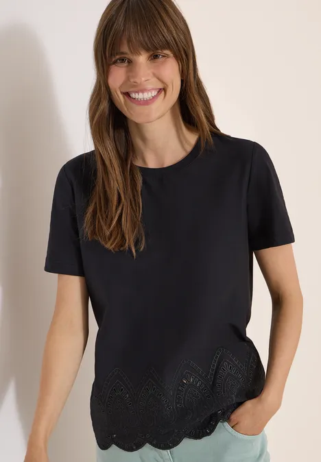 Een vrouw draagt een zwart T-shirt met korte mouwen en gezoomd ajourpatroon, hand in de spijkerbroekzak.