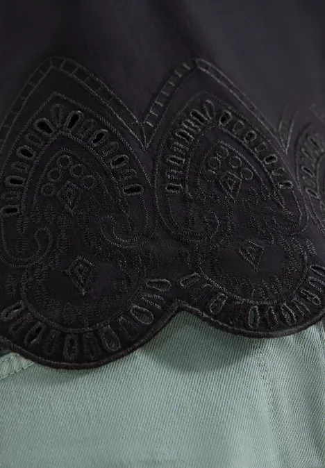 Zwarte stof met bloemrijke borduursels en gegolfde zoom, daaronder lichtgroen textiel.