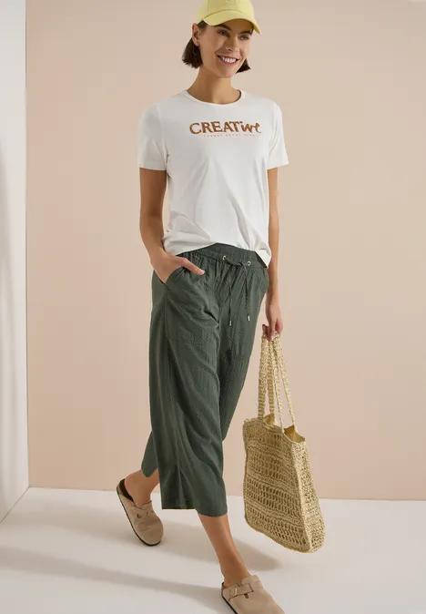 Frau trägt weißes T-Shirt mit 'CREATIVE'-Aufdruck, grüne Culotte, Strohtasche und beigen Mules vor neutralem Hintergrund.