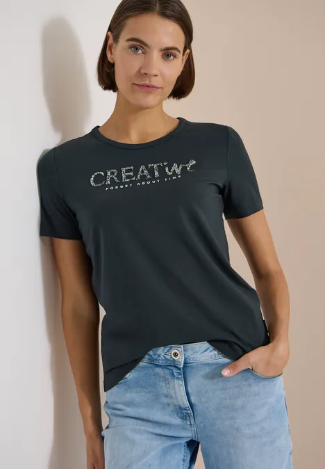 Frau trägt Jeans und dunkelgrünes T-Shirt mit Aufdruck 'CREATIV+', steht lässig vor neutralem Hintergrund.