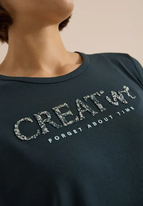 Frau trägt dunkelblaues T-Shirt mit glitzerndem Aufdruck 'CREATE UP' und Text 'FORGET ABOUT TIME'.