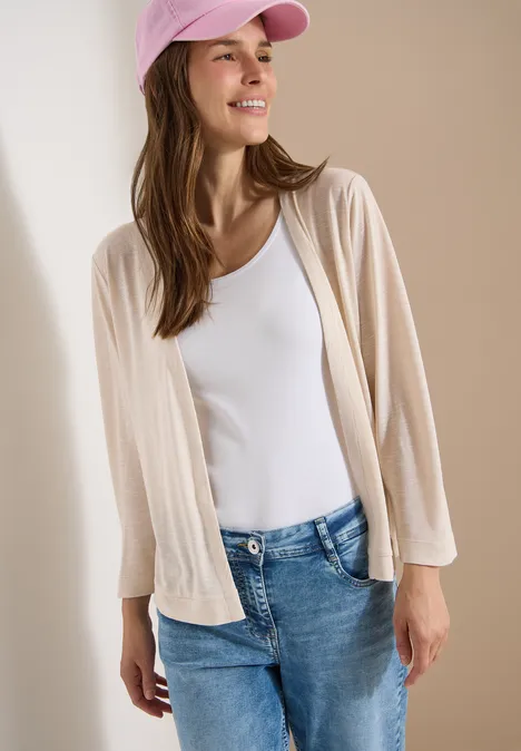 Vrouw met roze pet, wit topje, beige vest en spijkerbroek voor een neutrale achtergrond.
