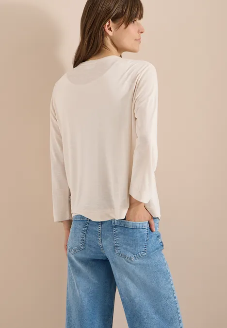 Persoon van achteren in een beige longsleeve en blauwe jeans, met één hand in de achterzak.