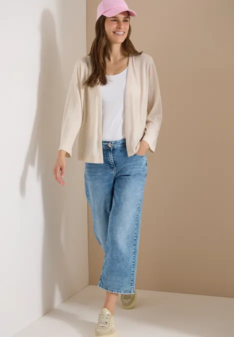 Vrouw tegen lichte muur, draagt wit topje, vest, blauwe jeans en lichte sneakers.
