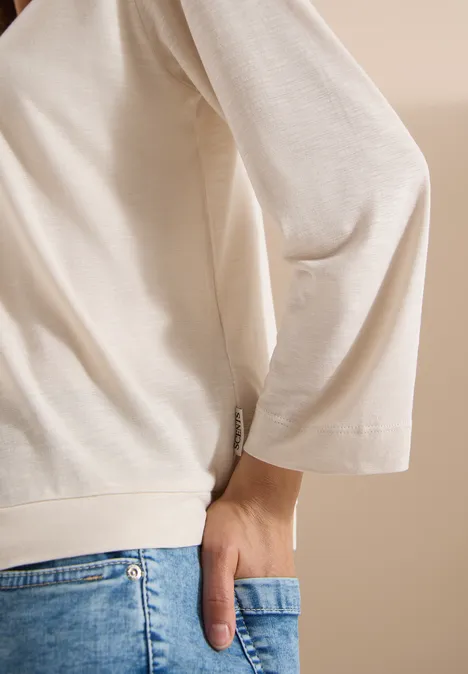 Persoon draagt crèmekleurig shirt en blauwe jeans, hand in achterzak.