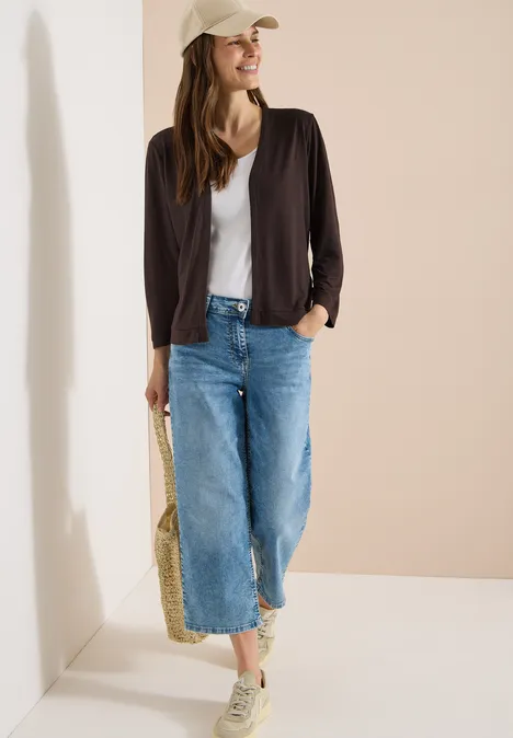 Vrouw in casual outfit: wit topje, zwarte jas, wijde jeans, sneakers, pet en rieten tas.