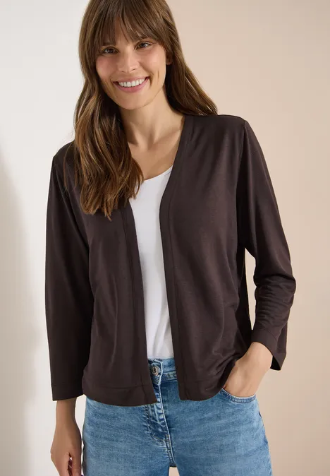 Vrouw met donkere vest over wit T-shirt en blauwe spijkerbroek, hand in zak.
