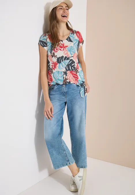 Frau lehnt an Wand, trägt bunt gemustertes T-Shirt, weite Jeans und weiße Sneakers.