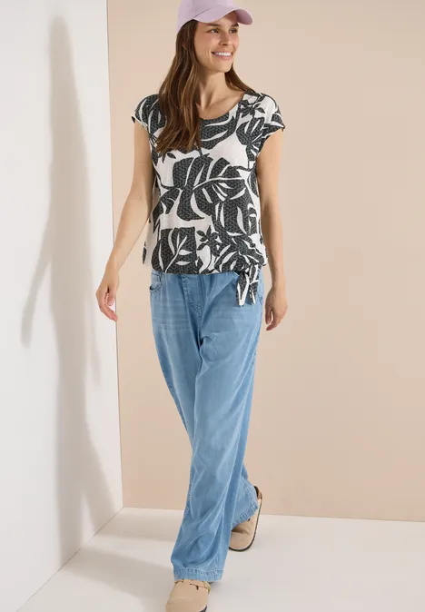T-Shirt in Ausbrenner-Optik palm breeze green Frau trägt ein kurzärmeliges Shirt mit schwarzem Blattmuster und weite hellblaue Jeans, steht in hellem Raum.