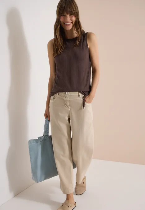 Vrouw in donkerbruin mouwloos shirt, beige wijd model broek en blauwe katoenen tas, voor lichte achtergrond.