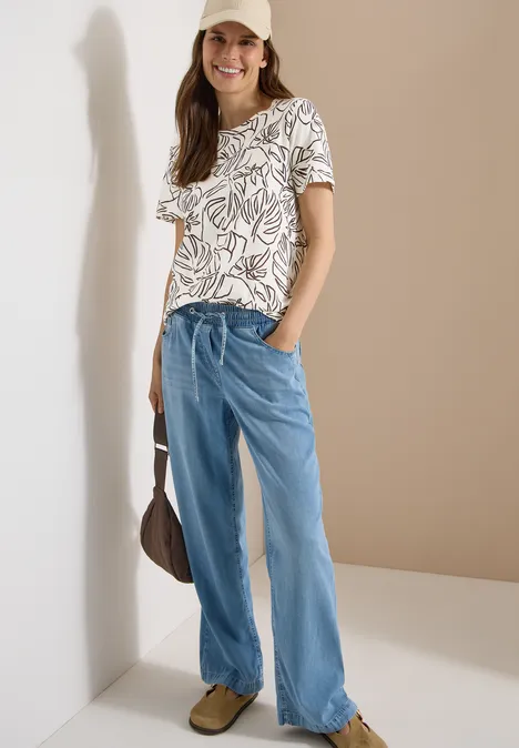 Vrouw met wit T-shirt met bladprint, wijde lichtblauwe jeans, bruine tas en schoenen.