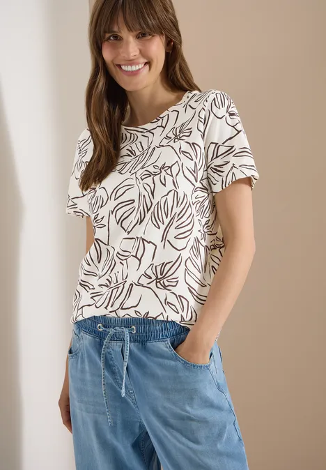 Vrouw in wit t-shirt met bladmotief en blauwe, losse broek voor een beige achtergrond.