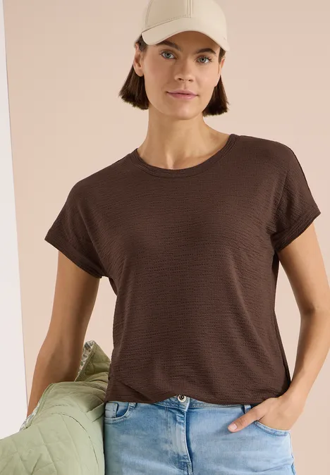 T-shirt à col rond et structure brown cacao Frau mit braunem T-Shirt und Jeans lehnt sich entspannt an grünes Kissen vor hellbeigem Hintergrund.