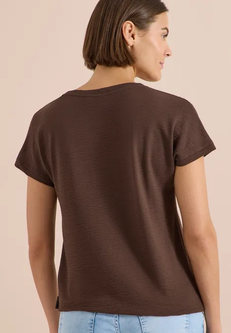 T-shirt à col rond et structure brown cacao Frau von hinten in braunem T-Shirt und Jeans vor beigem Hintergrund.