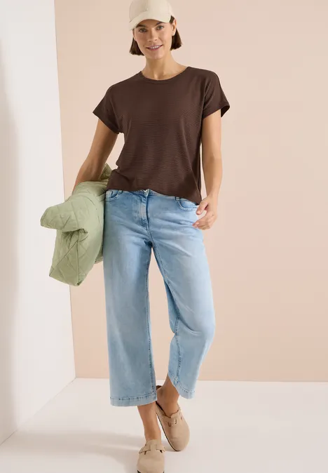 T-shirt à col rond et structure brown cacao Modell in braunem T-Shirt, hellen Jeans und beigen Schuhen hält eine grüne Jacke in der Hand.