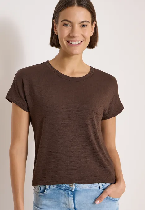 T-shirt à col rond et structure brown cacao Frau trägt braunes T-Shirt und blaue Jeans, Hand in Hosentasche, vor hellem Hintergrund.