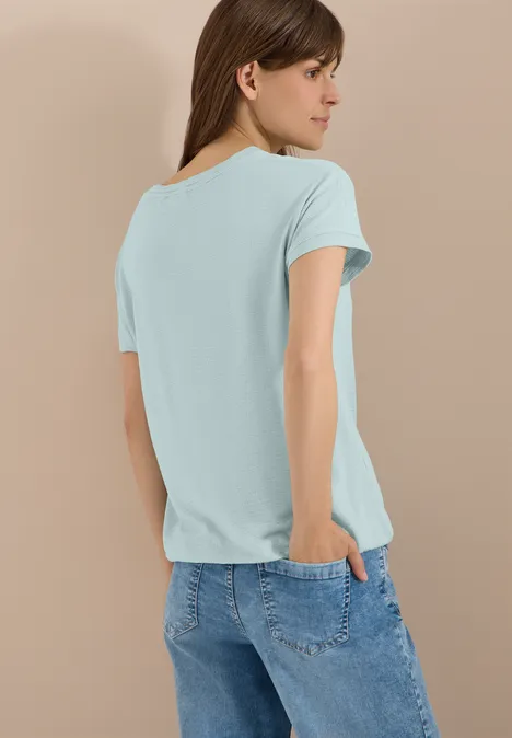Shirt met ronde hals en structuur crystal mint Vrouw van achter in turquoise T-shirt en blauwe jeans voor een beige achtergrond.