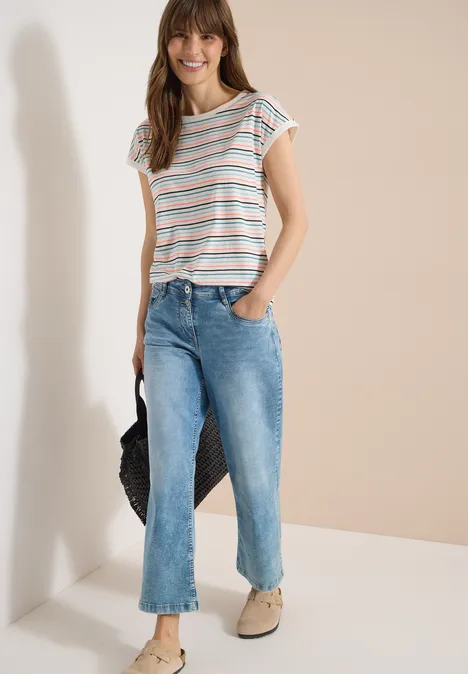 T-Shirt mit Boat Neck und Turn-Up-Detail vanilla white Frau lehnt an heller Wand in gestreiftem T-Shirt, blauen Jeans, Tragekorb und beigen Slippern.