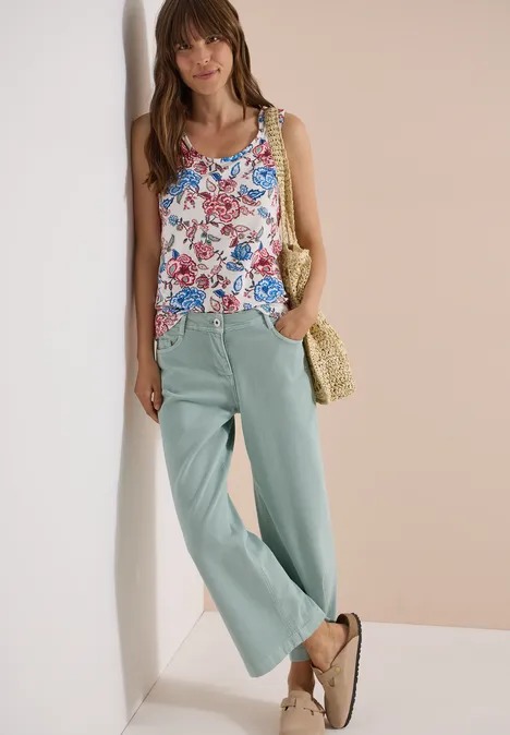 Frau lehnt lässig an Wand, trägt florales Top, helle weite Hose, Strohtasche und Sandalen.