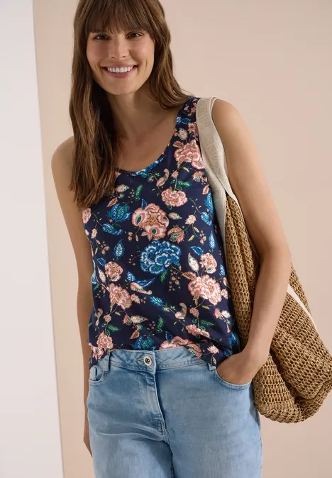Frau in einem navyblauen, geblümten Top und Jeans trägt eine geflochtene Strohtasche.