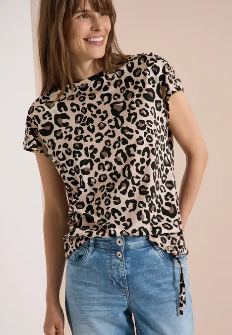 T-Shirt mit Raffung und Print urban taupe Frau in Leopardenmuster-T-Shirt mit seitlichen Bändern kombiniert mit blauer Jeans vor neutralem Hintergrund.