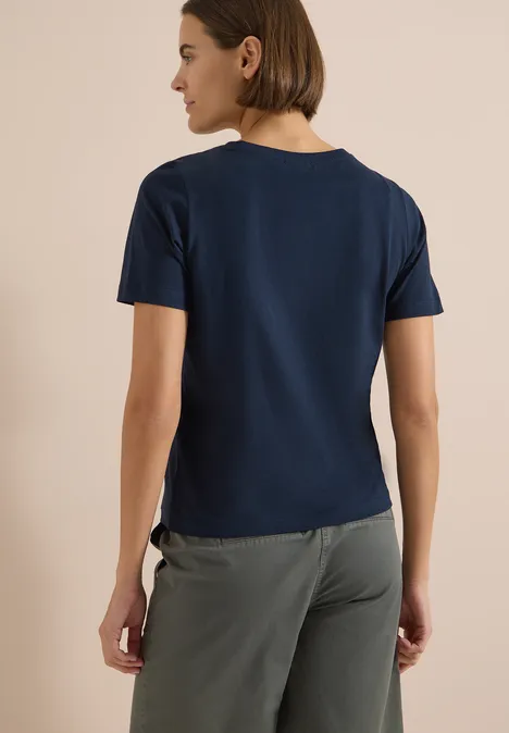 T-Shirt mit Boat Neck und Wording Artwork universal blue Frau von hinten in dunkelblauem T-Shirt und grauer Hose vor hellem Hintergrund.