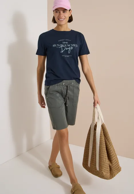 T-Shirt mit Boat Neck und Wording Artwork universal blue Eine Person trägt ein navy T-Shirt, graue Shorts, braune Mokassins und hält eine große geflochtene Tasche.
