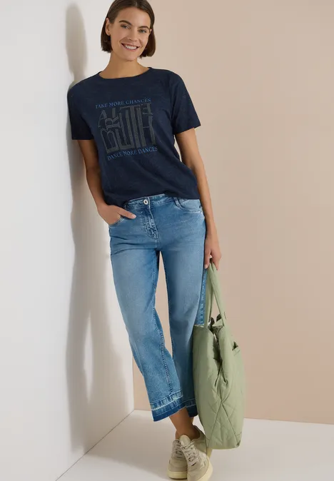 T-Shirt in Ausbrenner-Optik universal blue Frau lehnt lässig an der Wand in dunkelblauem T-Shirt, hellen Jeans und hält eine grüne Tasche.