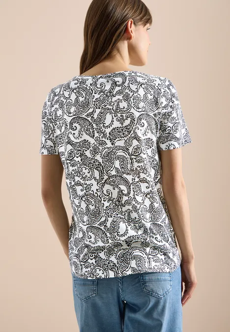Vrouw van achter gefotografeerd in een wit T-shirt met zwart paisley-patroon en een blauwe jeans.
