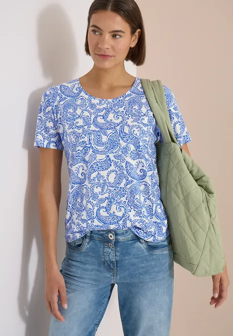 Vrouw draagt blauw-wit geprint T-shirt, jeans en groene gewatteerde tas.