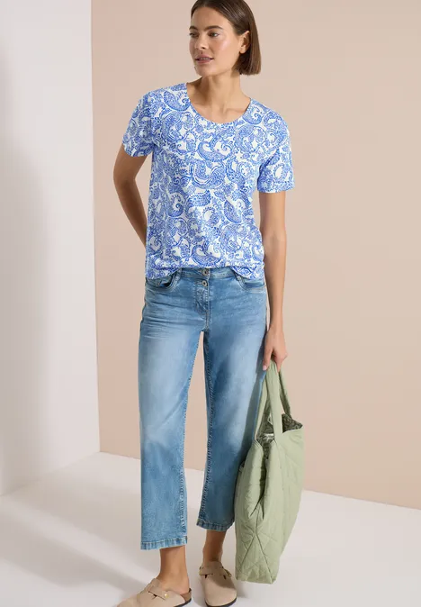 Vrouw met blauw print-T-shirt, lichte spijkerbroek, groene tas en sandalen voor een beige achtergrond.