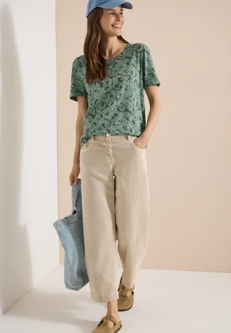 Frau trägt grün gemustertes T-Shirt, beige Hose, blaue Tasche und Cap.