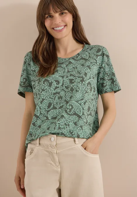 Frau trägt olivgrünes, gemustertes T-Shirt und beige Hose, Hände in den Taschen.