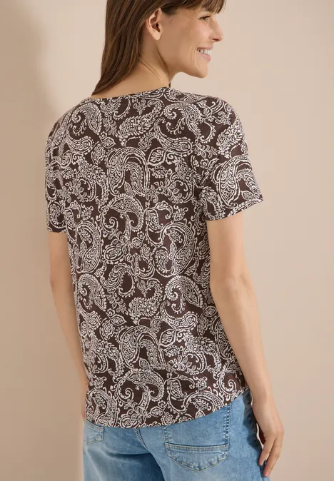 Rückenansicht einer Frau in T-Shirt mit Paisley-Muster und Jeans vor neutralem Hintergrund.
