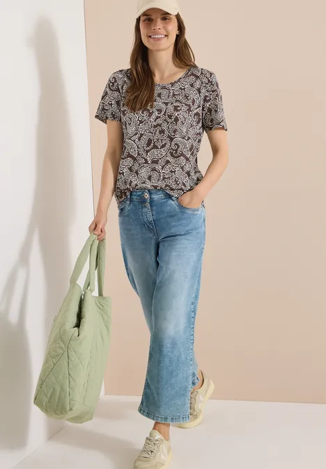 Eine Frau trägt ein gemustertes T-Shirt, weite Jeans und eine grüne Stepp-Tasche vor beigem Hintergrund.