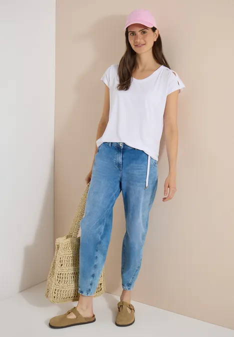 Jonge vrouw in casual zomerse outfit: wit t-shirt, ruime jeans, sandalen, roze pet en net tas.