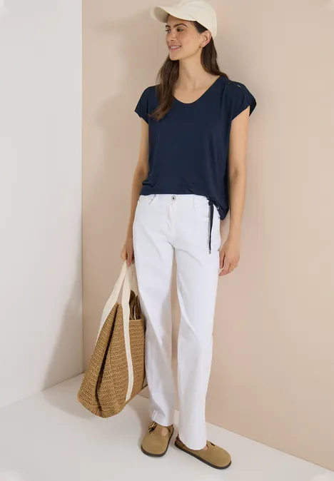 Vrouw in marineblauw T-shirt, witte broek en witte pet, met strooien tas en beige loafers voor een beige muur.