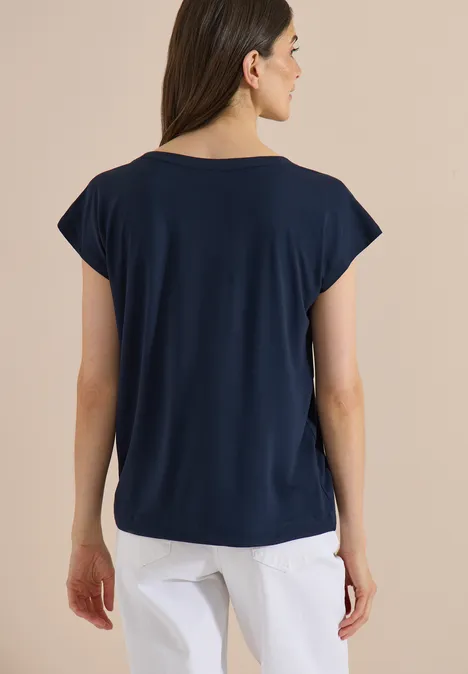 Een vrouw van achteren draagt een donkerblauw T-shirt met korte mouwen en een witte broek voor een neutrale achtergrond.