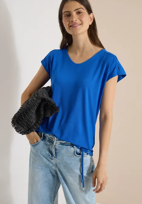 Vrouw in blauw T-shirt met trekkoord, lichte jeans en gestructureerde zwarte tas over de schouder, lichte achtergrond.