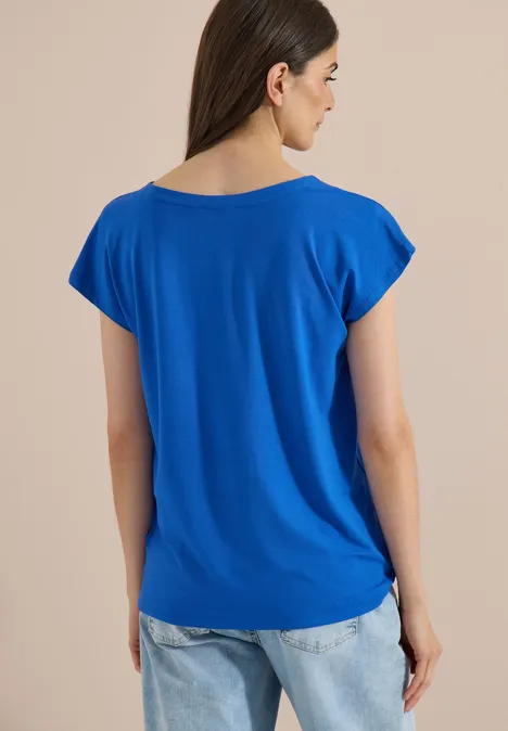 Vrouw van achteren in een losse blauwe T-shirt en lichtblauwe spijkerbroek tegen een neutrale achtergrond.