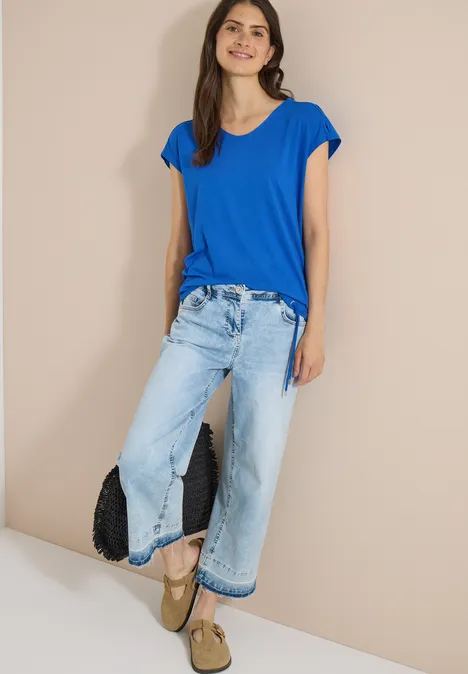 Vrouw in blauwe T-shirt, wijde jeans, plateauzolen en zwarte clutch voor een beige achtergrond.
