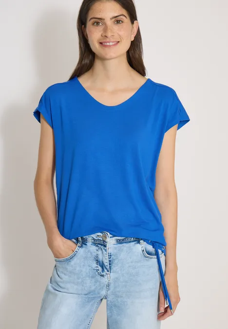 Vrouw in blauw V-hals T-shirt en lichtblauwe spijkerbroek met handen in de zakken, lichte achtergrond.