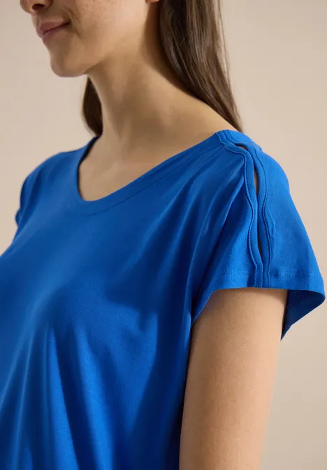 Een vrouw in profiel draagt een felblauw T-shirt met ronde hals en licht geplooide korte mouwen.