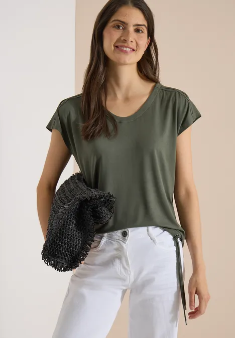 Eine Frau trägt ein grünes T-Shirt und weiße Jeans, hält einen schwarzen Netzbeutel vor neutralem Hintergrund.