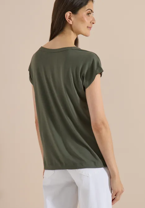 Rückenansicht einer Frau in grünem T-Shirt mit umgeschlagenen Ärmeln und weißer Hose vor hellem Hintergrund.