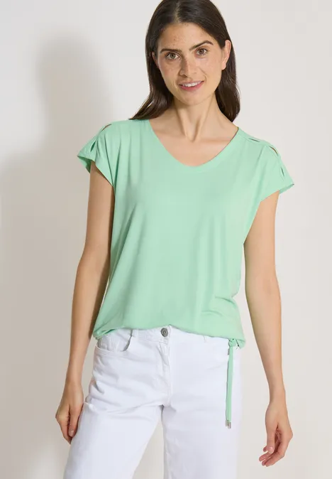 Model draagt lichtgroen T-shirt met korte mouwen en witte broek tegen een effen achtergrond.