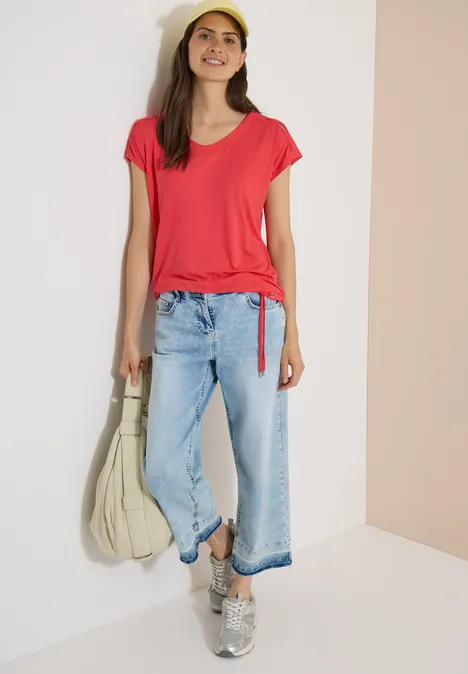 Vrouw in rood T-shirt, lichtblauwe jeans en sneakers, met grote tas, tegen een neutrale achtergrond.