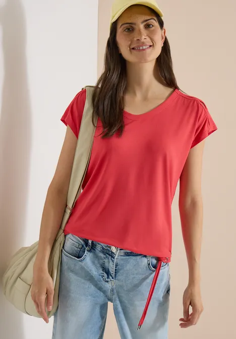 Vrouw in een rood t-shirt, spijkerbroek en schoudertas voor een lichte achtergrond.