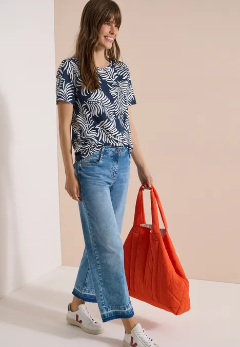 Vrouw draagt blauw t-shirt met bladerenprint, wijde cropped jeans, witte plateau-espadrilles en grote rode tas.